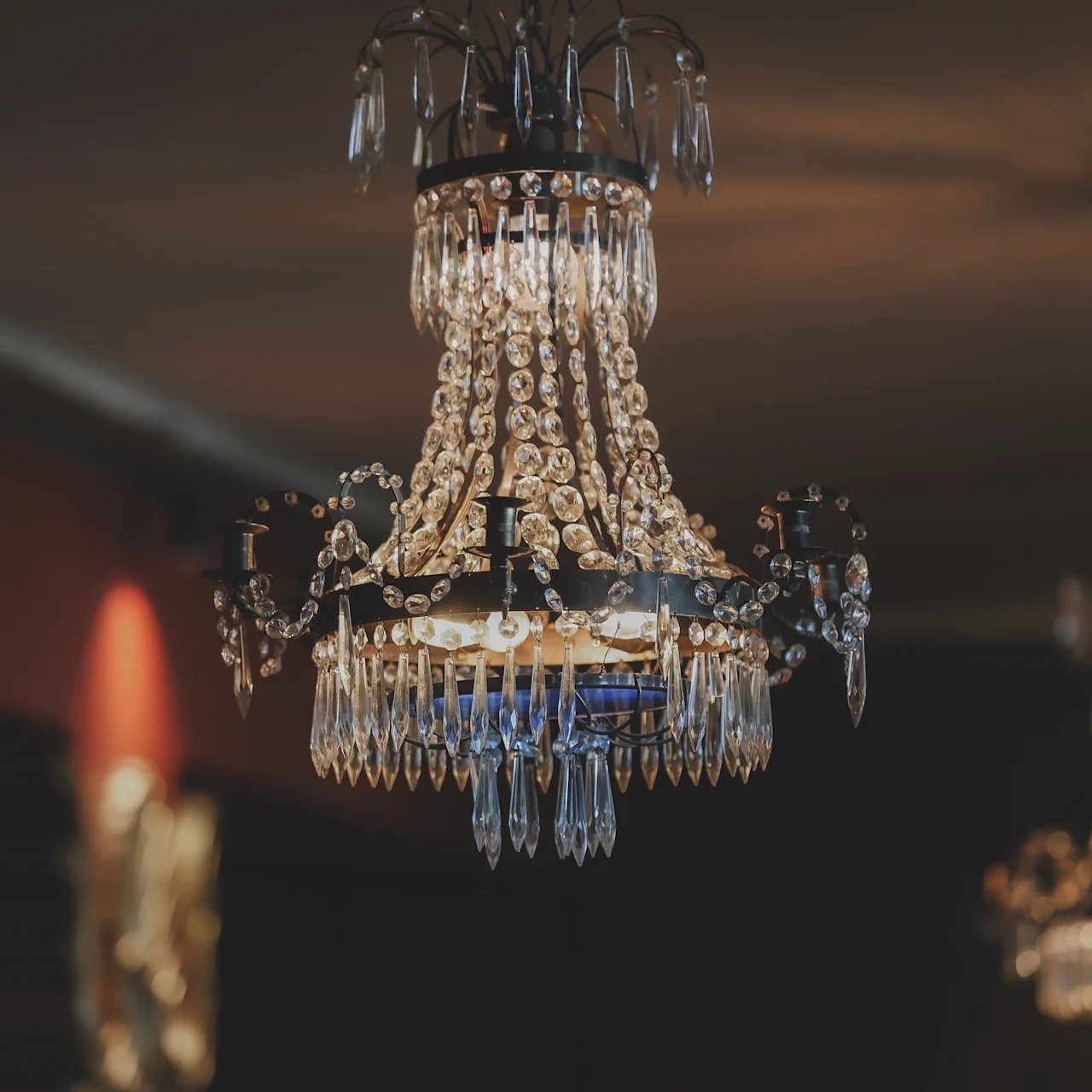 A crystal chandelier hangs from the ceiling, casting a warm glow in a dimly lit, elegant room, with another chandelier visible in the background.