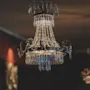 A crystal chandelier hangs from the ceiling, casting a warm glow in a dimly lit, elegant room, with another chandelier visible in the background.