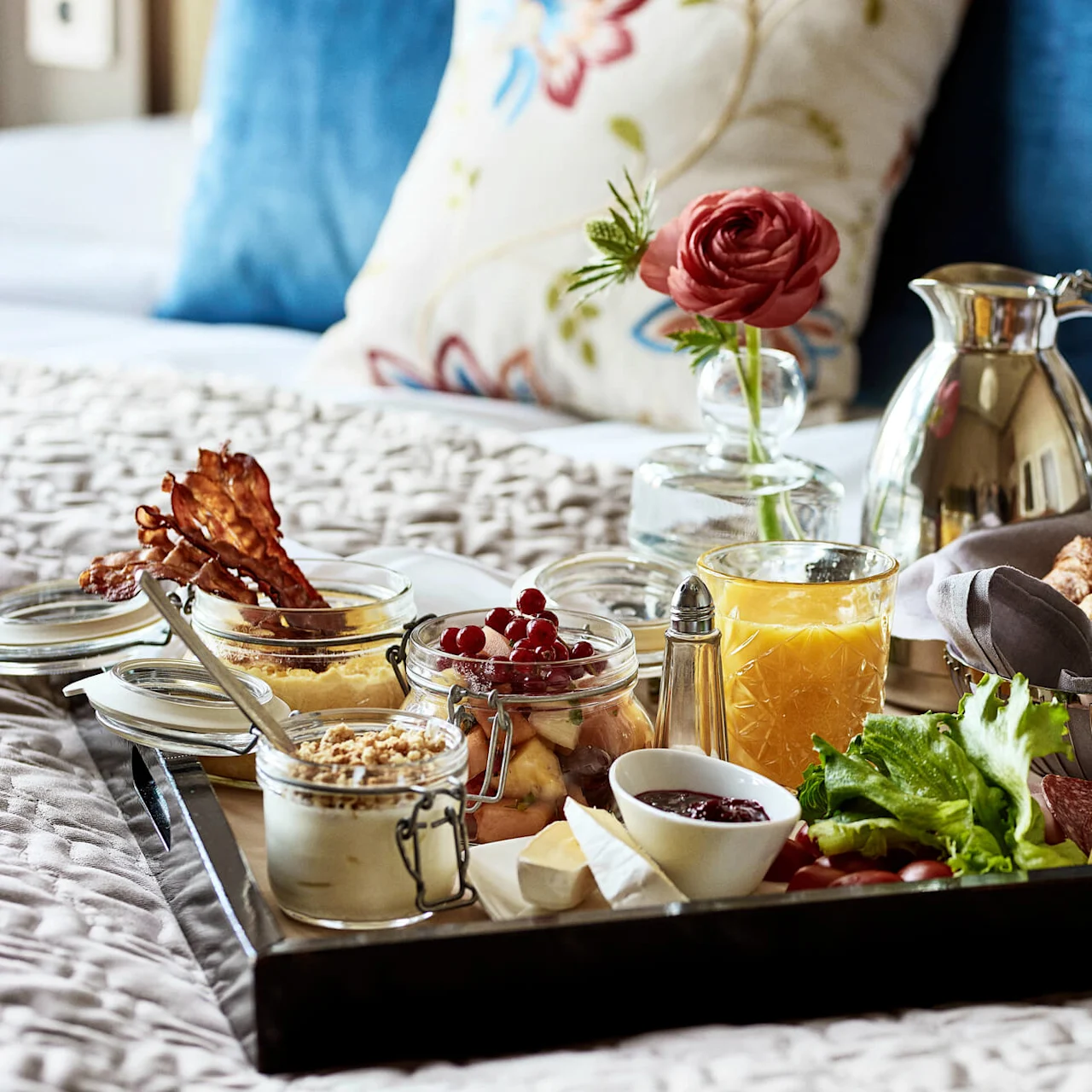A tray with croissants, fruit, bacon, cheese, a flower vase, and a glass of orange juice sits on a bed. Decorative pillows and a blanket provide a cozy background.