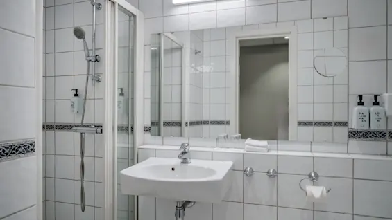 Et lyst hotellbad med dusj, vask og speil. Fasiliteter inkluderer håndsåpe og lotion.