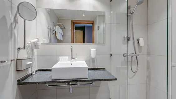 A modern bathroom features a white sink on a dark countertop, with a wall-mounted mirror above. A glass-enclosed shower with a handheld spray is on the right, surrounded by white tiles.