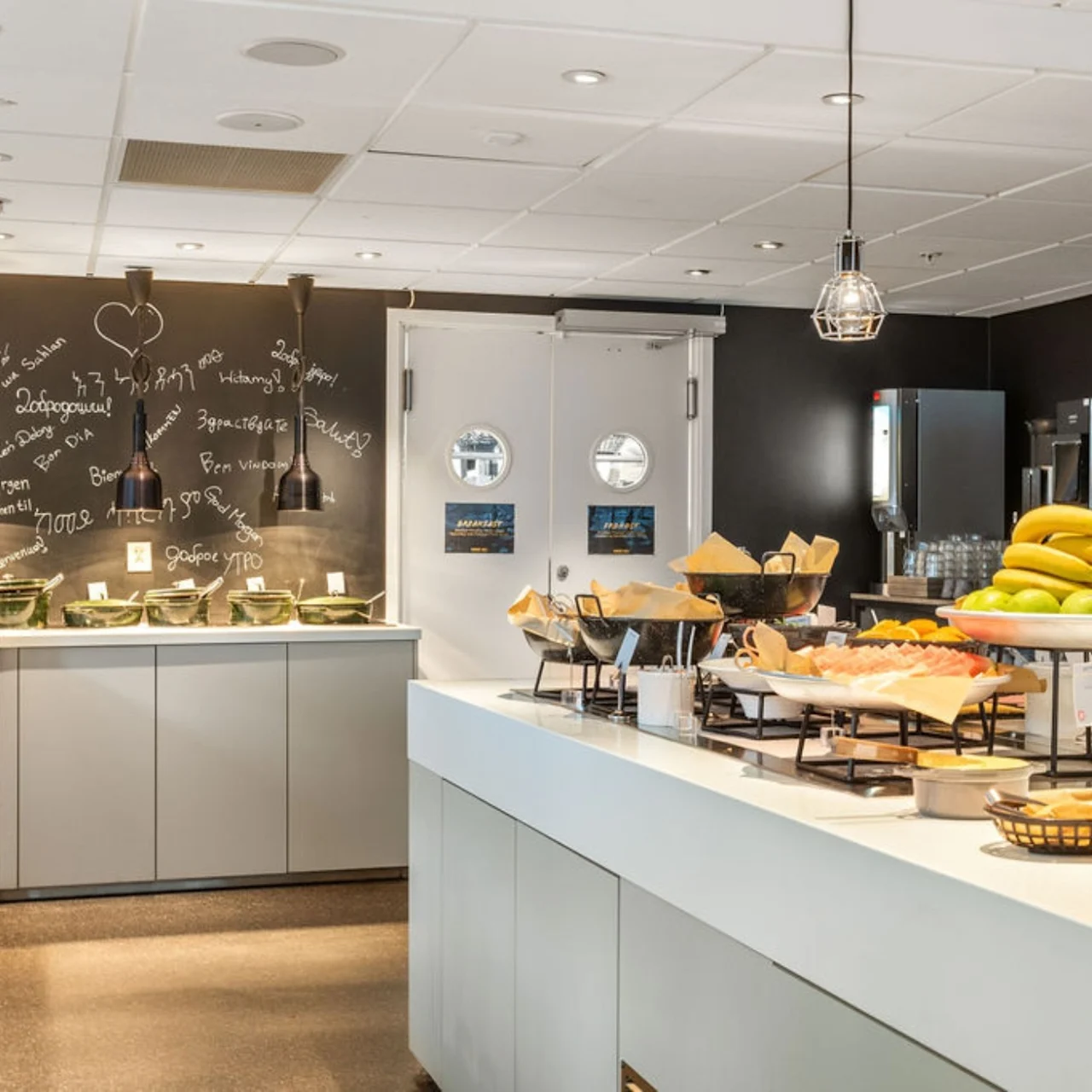 Buffet counter displaying bananas, bread, and pastries in a spacious room with a chalkboard wall featuring written notes and a heart shape. Ceiling lamps provide lighting.