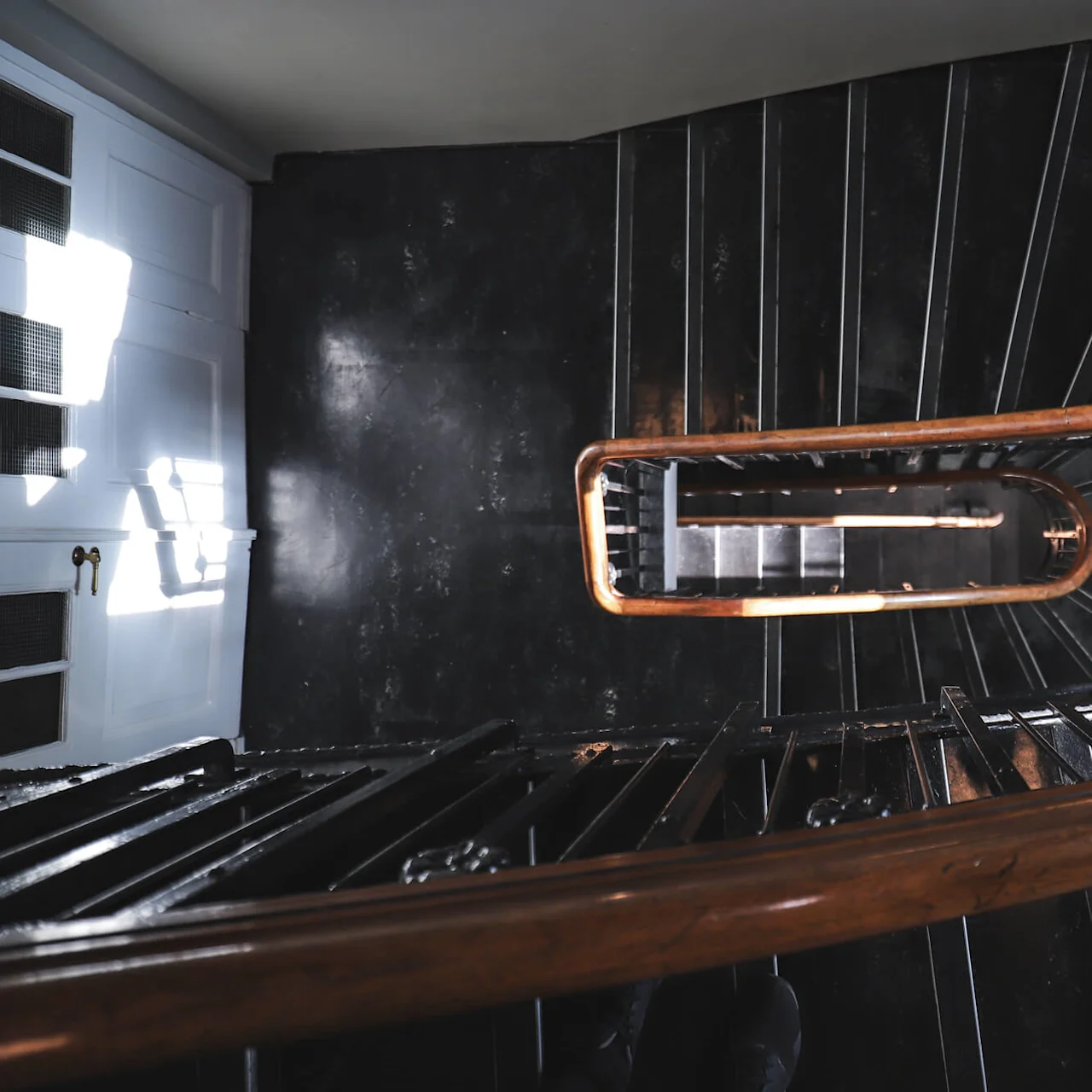 A wooden handrail spirals downward, encircling a staircase with dark steps in a dimly lit building. Sunlight filters through a nearby window, casting shadows on the wall.
