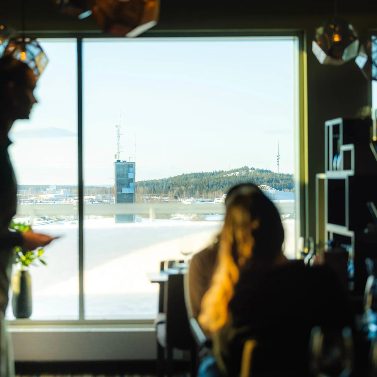 Silhuett av en person som går inomhus nära ett stort fönster, utomhus syns ett snötäckt landskap, träd och ett torn i dagsljus.