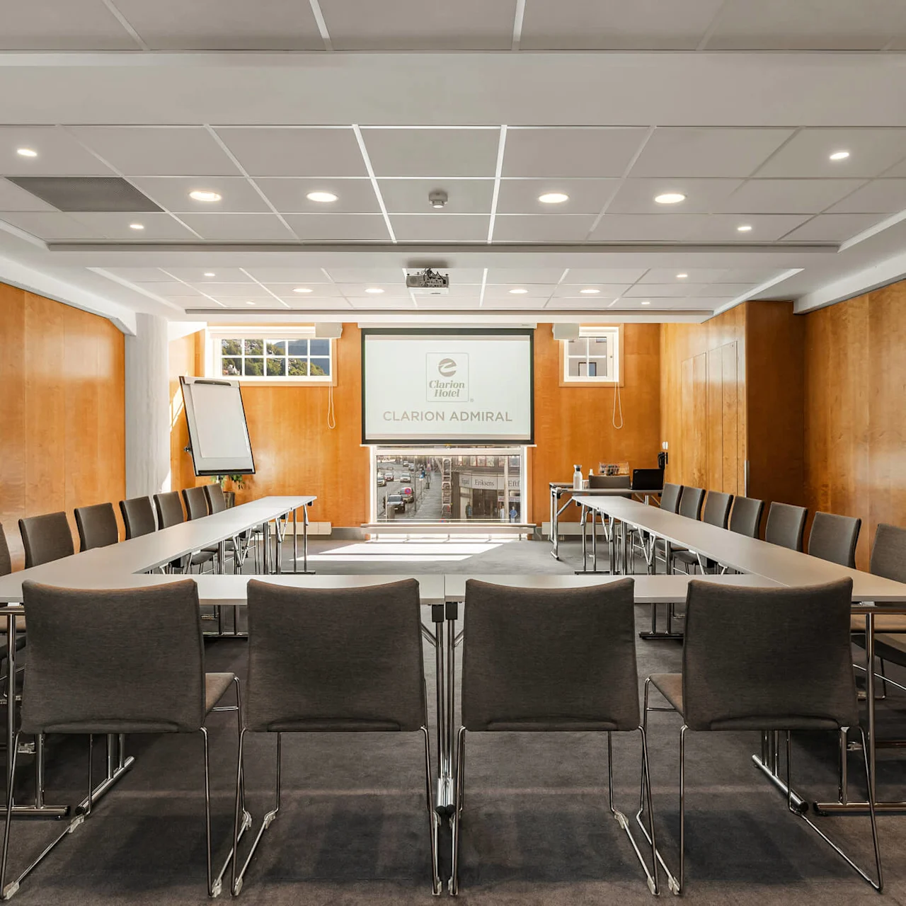 Konferencerum med U-formet bord arrangement omgivet af stole, vendt mod en projektorskærm, der viser "Clarion Hotel CLARION ADMIRAL." Lyse træpaneler og loftslamper.