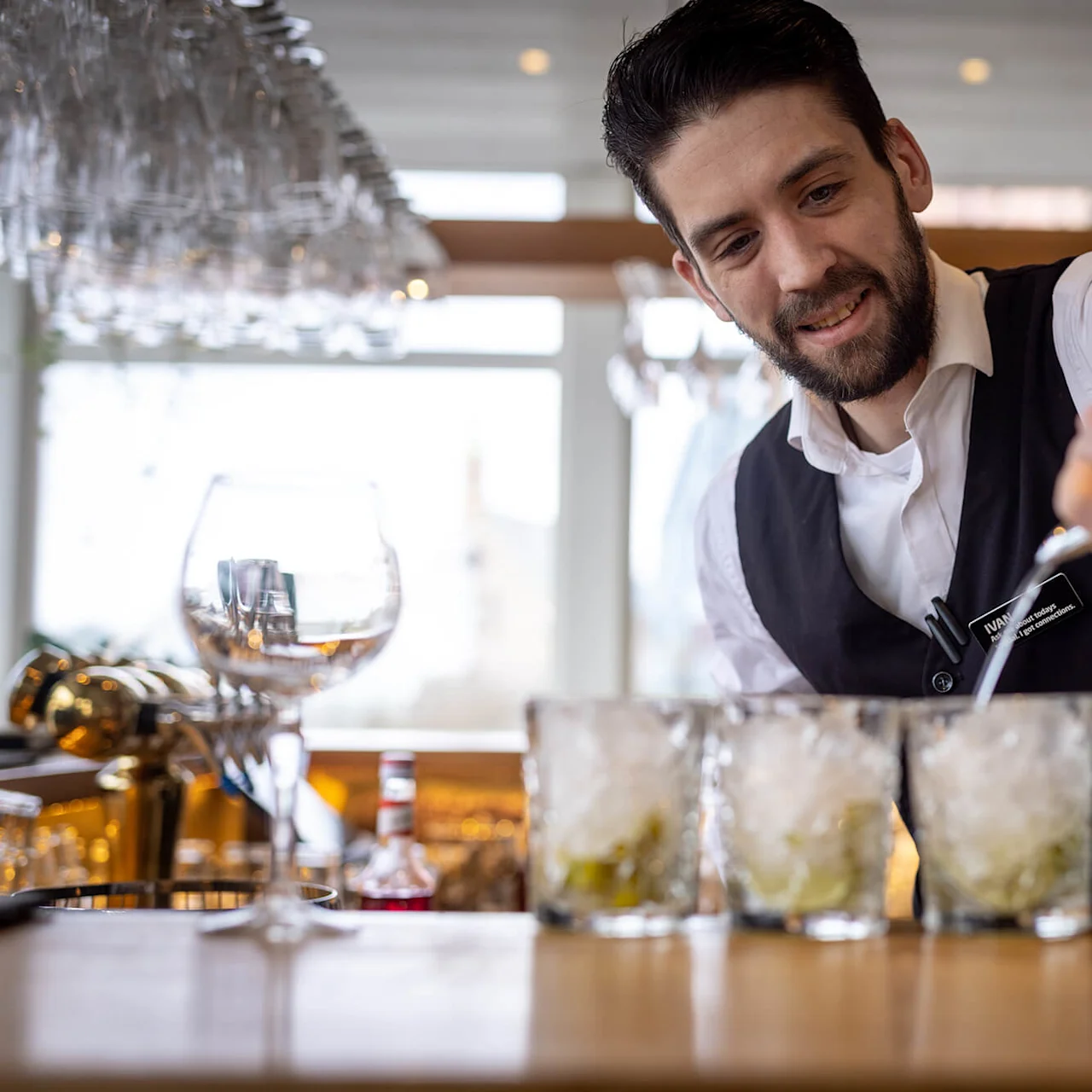 Bartender pours clear liquid into four glasses filled with ice on a counter, surrounded by a bar setting with hanging glasses. Name tag reads "NAME, Bar Manager."