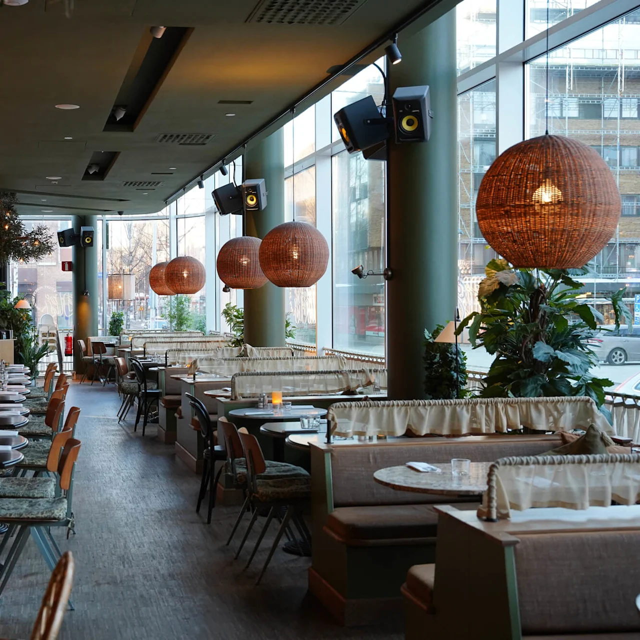 En moderne restaurantinteriør med træborde, behagelige siddepladser, store vinduer og dekorative belysningsarmaturer.