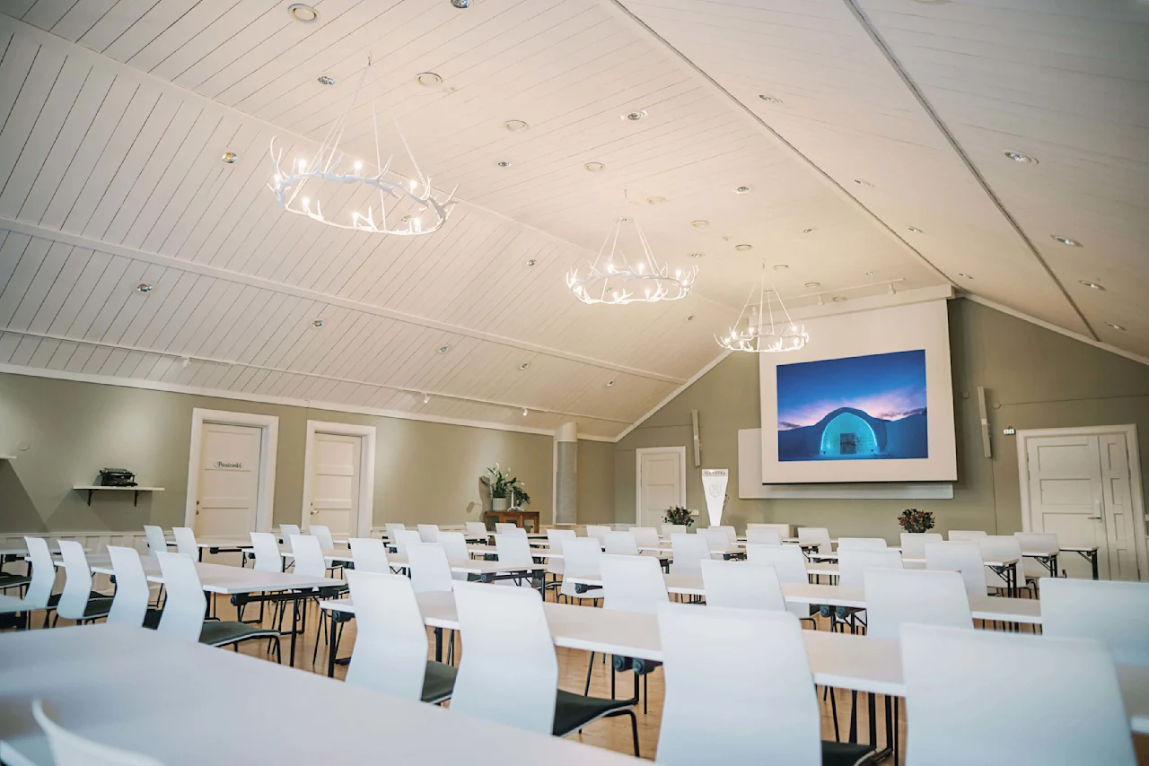 Rækker af hvide stole vender mod en stor skærm, der viser en blå isstruktur, placeret i et rummeligt rum med et hvælvet loft og kronformede lysekroner. En dør mærket "Porjuseli" er synlig.