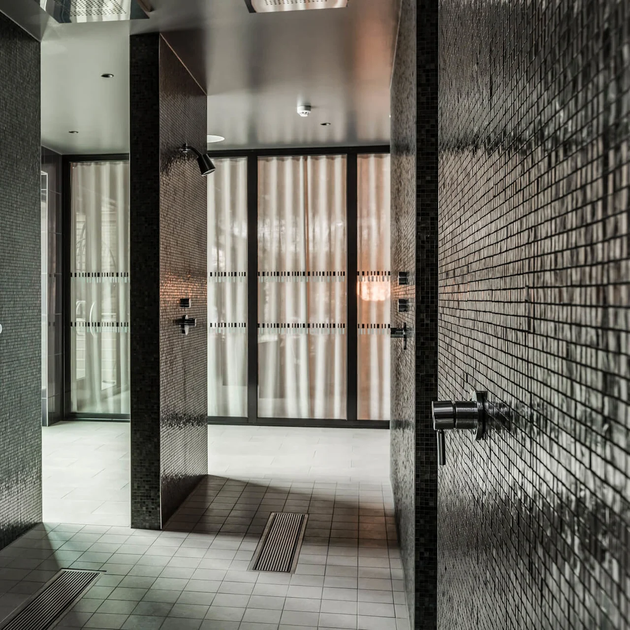 Brusere med mørke mosaikfliser står ubrugte i et moderne badeværelse. Blødt lys filtreres gennem matteret glas, hvilket skaber en rolig stemning.
