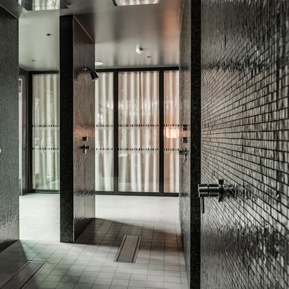 Showers with dark mosaic tiles stand unused in a modern bathroom. Soft light filters through frosted glass doors, creating a serene atmosphere.