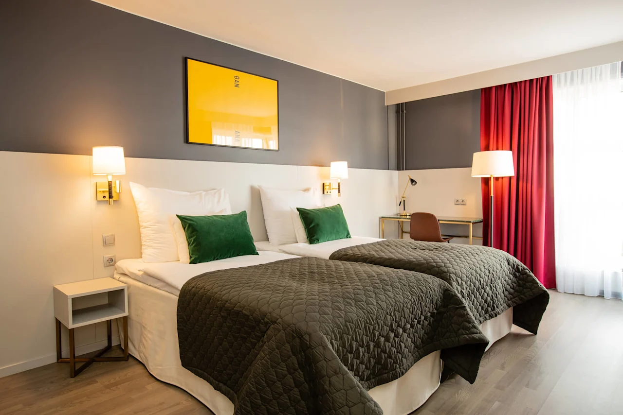 Kaksi siististi pedattua erillistä sänkyä vihreillä tyynyillä ja harmailla peitteillä sijaitsevat modernissa hotellihuoneessa. Keltainen teos, jossa lukee "BANANA", roikkuu yllä; pöytä ja tuoli näkyvät punaisten verhojen lähellä.