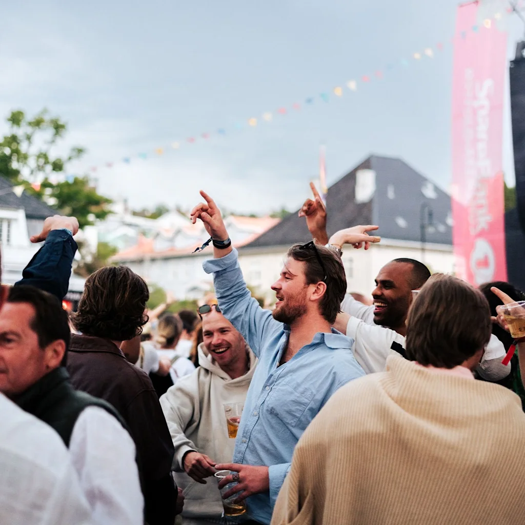 En livlig folkemengde nyter et utendørs arrangement, mange med hendene i været. Et banner med "Spenn ink" er synlig.