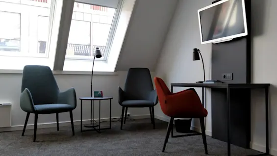 Et moderne kontorhjørne med tre stole omkring et lille bord under skrå vinduer, med et vægmonteret fjernsyn og en skrivebordslampe på siden.