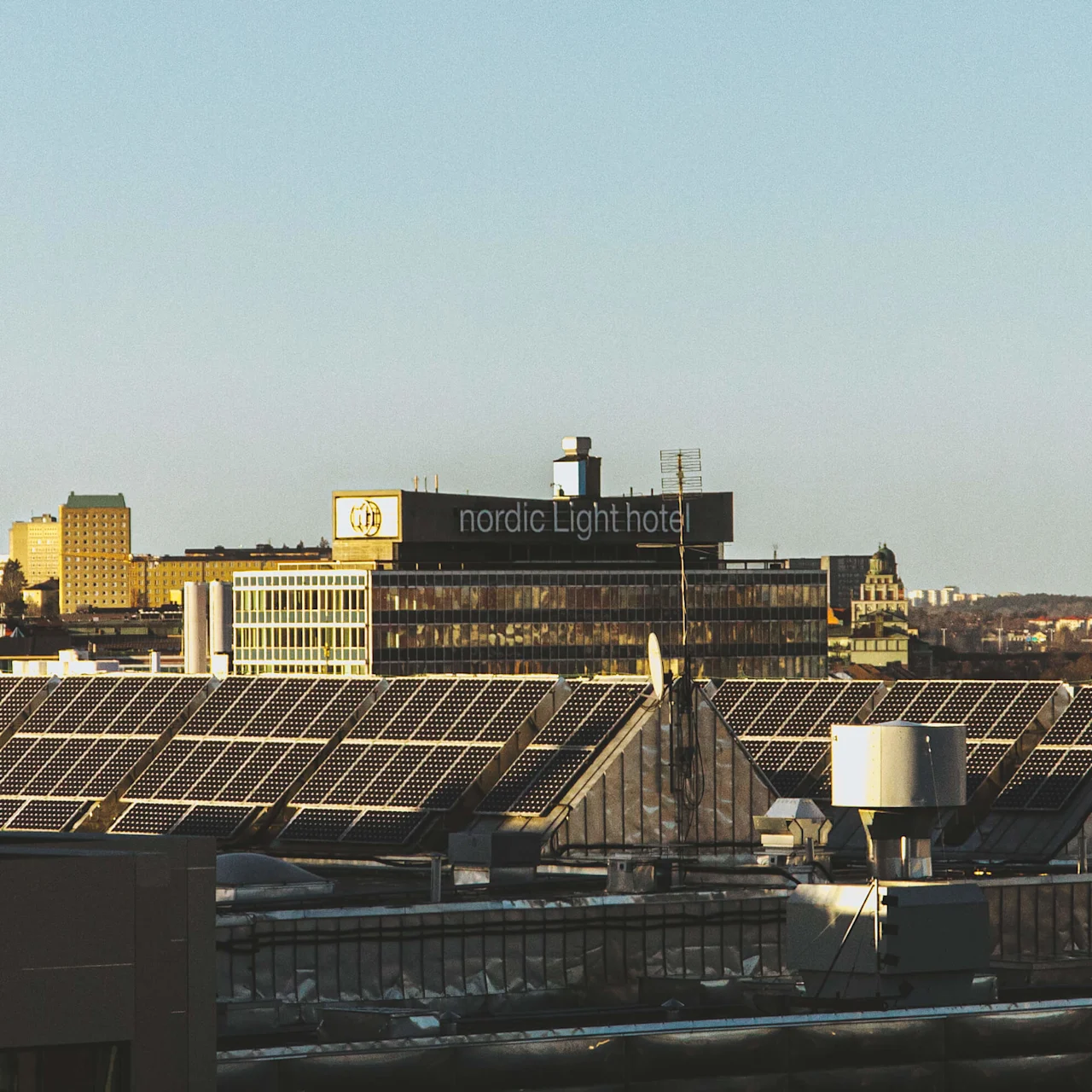 Solar panels are installed on a rooftop, reflecting sunlight. In the background, a building displays the signage "Nordic Light Hotel" surrounded by cityscape and clear blue sky.