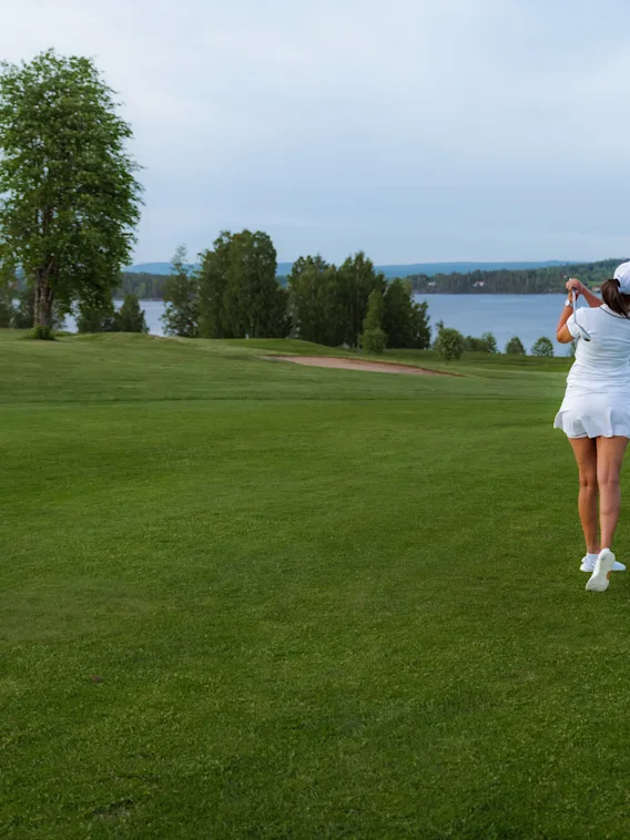 En golfspiller i hvitt antrekk svinger en kølle på en gressbane nær en innsjø, omgitt av trær under en skyet himmel.