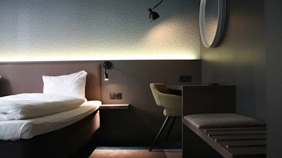 A neatly made bed with white linens stands in a modern, dimly lit hotel room featuring a desk, chair, and a wall lamp. A round mirror decorates the wall.