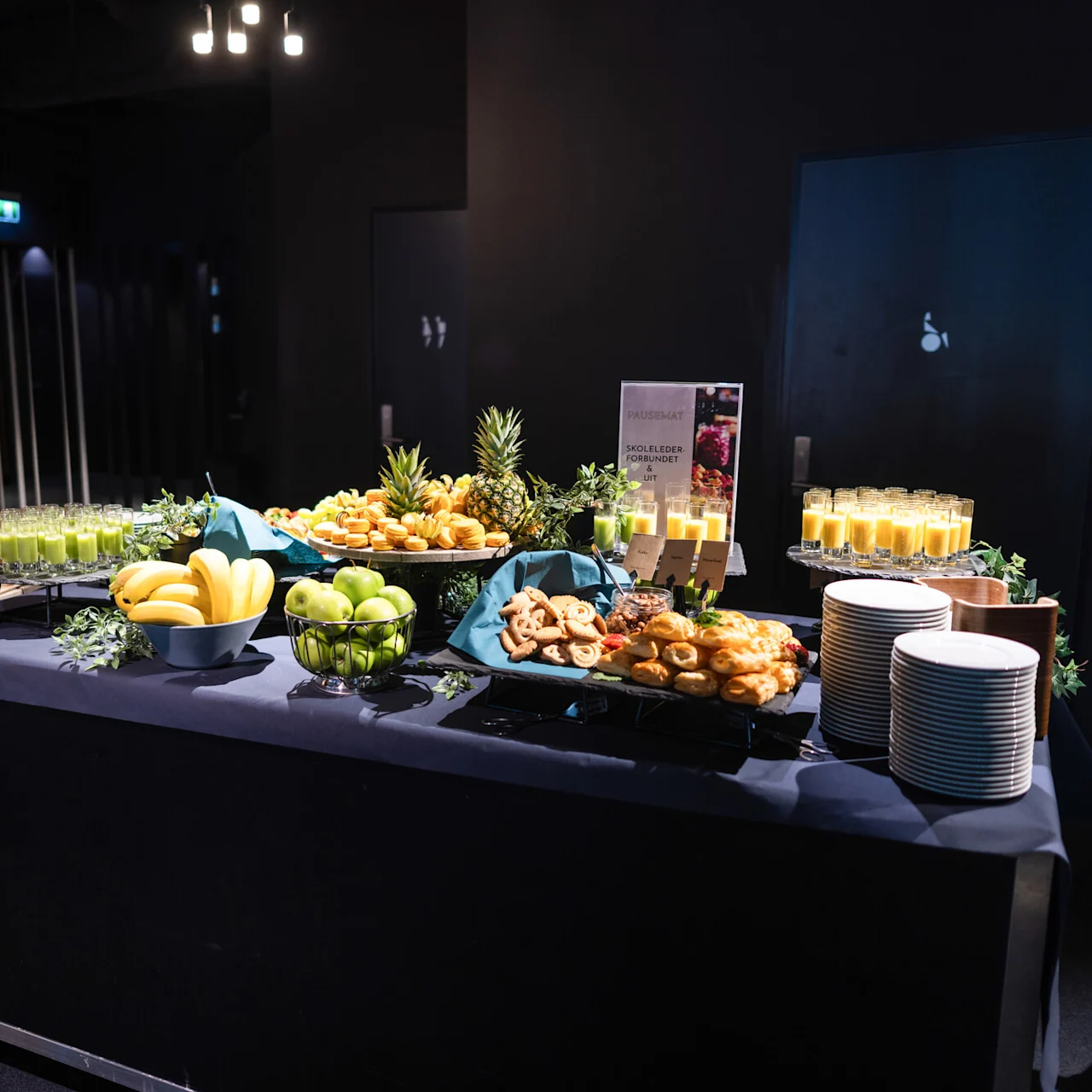 Et buffébord har fruktskåler, bakverk og juiceglass under varm belysning i en moderne innredning. Tekst på et skilt lyder "PAUSESMART SKOLELEDERFORBUNDET." Stablede tallerkener er klare i nærheten.