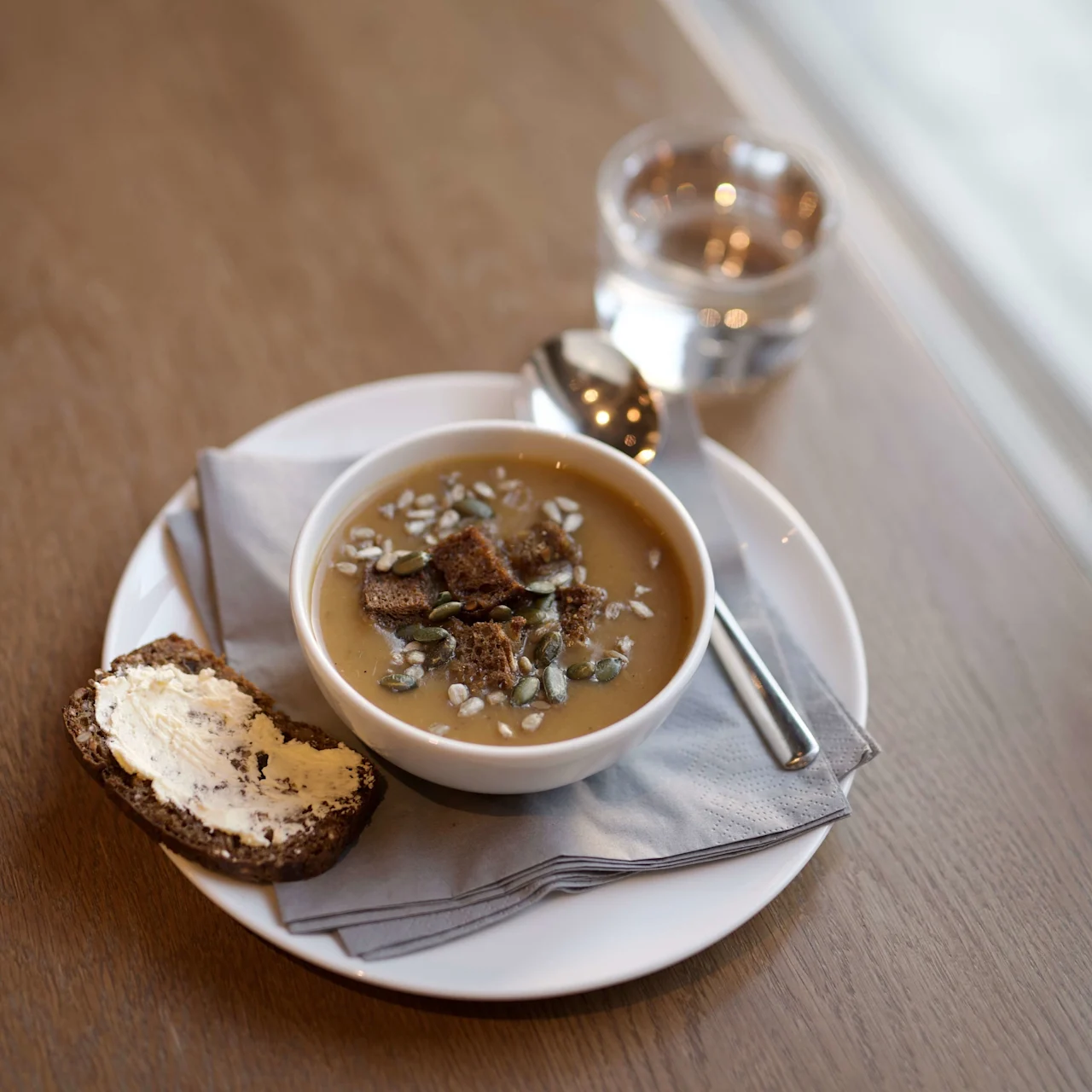 A bowl of soup with seeds and croutons, a slice of bread with butter; a meal on a wooden table beside a window.