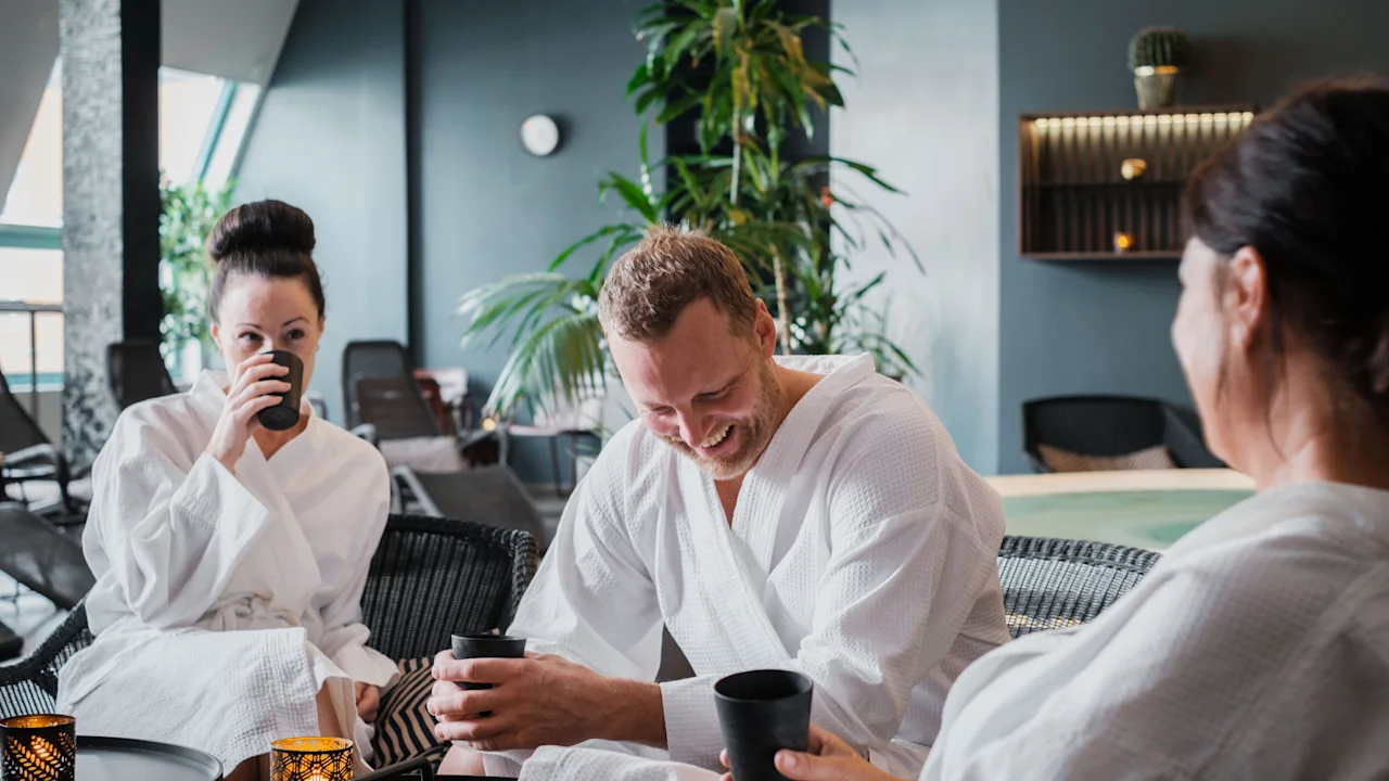 Tre personer i badrockar kopplar av i en lounge, njuter av drycker och samtal. En person skrattar.