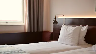 En säng med vita kuddar och en lampa på en trähuvudgavel i ett hotellrum bredvid ett fönster med gardiner.
