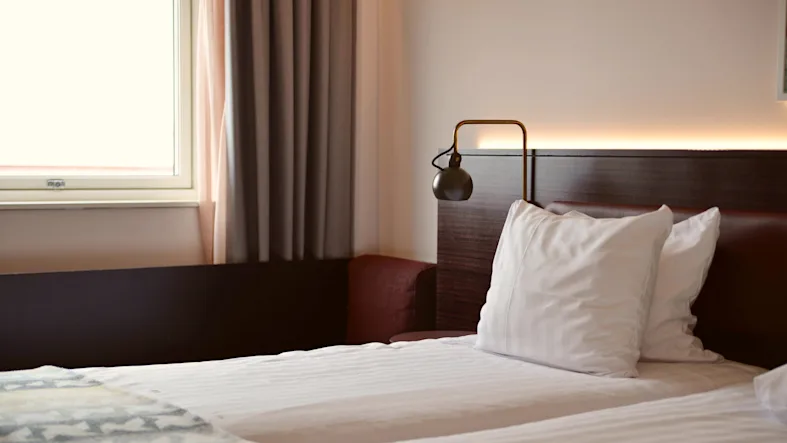A bed with white pillows and a lamp on a wooden headboard is in a hotel room next to a window with curtains.