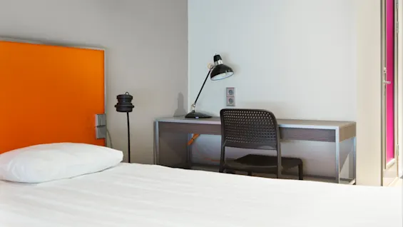 En seng med orange sengegavl står ved siden af et skrivebord med sort lampe og perforeret stol, under en loftslampe i et moderne, lyst rum.