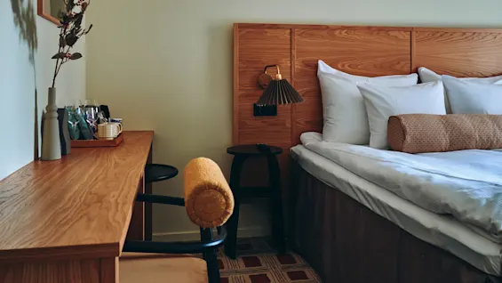 Et koselig hotellrom med en komfortabel seng, sengegavl i tre, et funksjonelt skrivebord og et mønstret teppe.