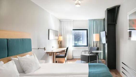 A neatly made bed with teal accents is positioned in a modern hotel room featuring a desk, armchair, and window with curtain, illuminated by ceiling and floor lamps.