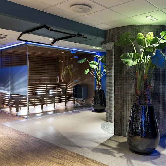 A spa area features large potted plants standing beside a glass-walled sauna. Warms lights illuminate the wooden interior, and towels are neatly stacked in the background.