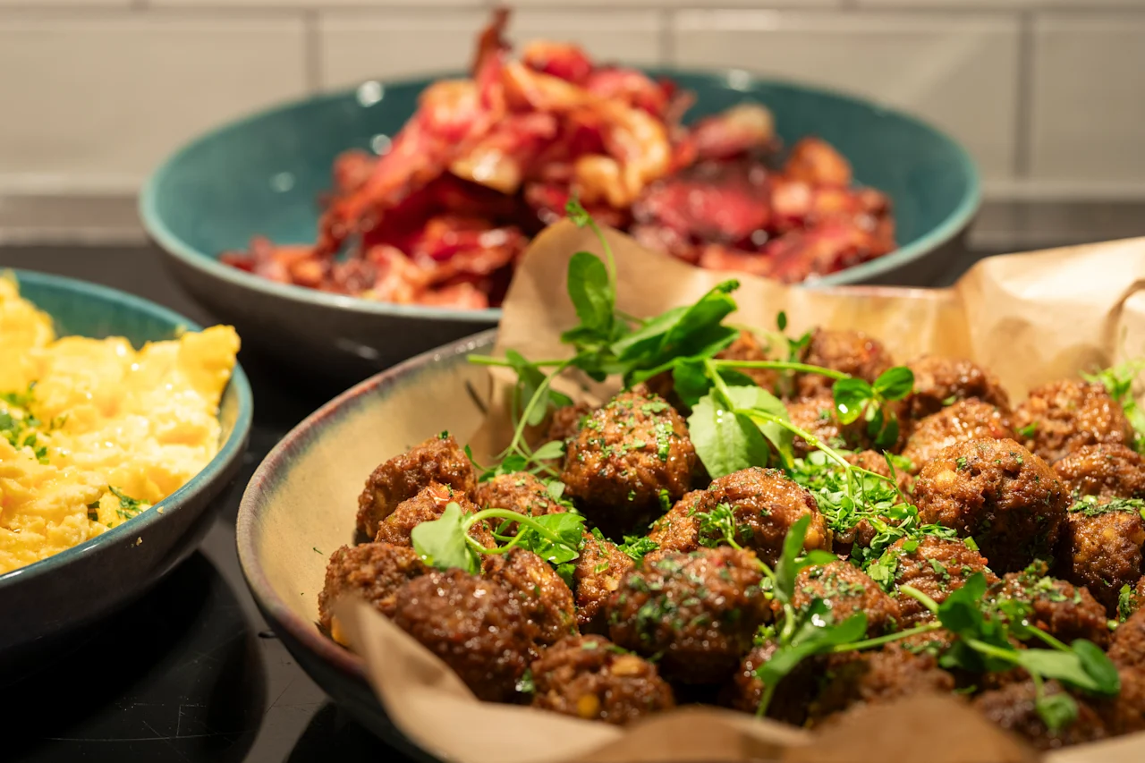 Krispiga köttbullar garnerade med färska örter och ruccola fyller en skål, omgivna av tallrikar med äggröra och krispig bacon på en mörk köksbänk.