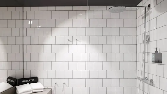 A modern bathroom with white tiled walls and a sleek shower fixture. Towels and a labeled "HAIRDRYER" case rest on a counter. 