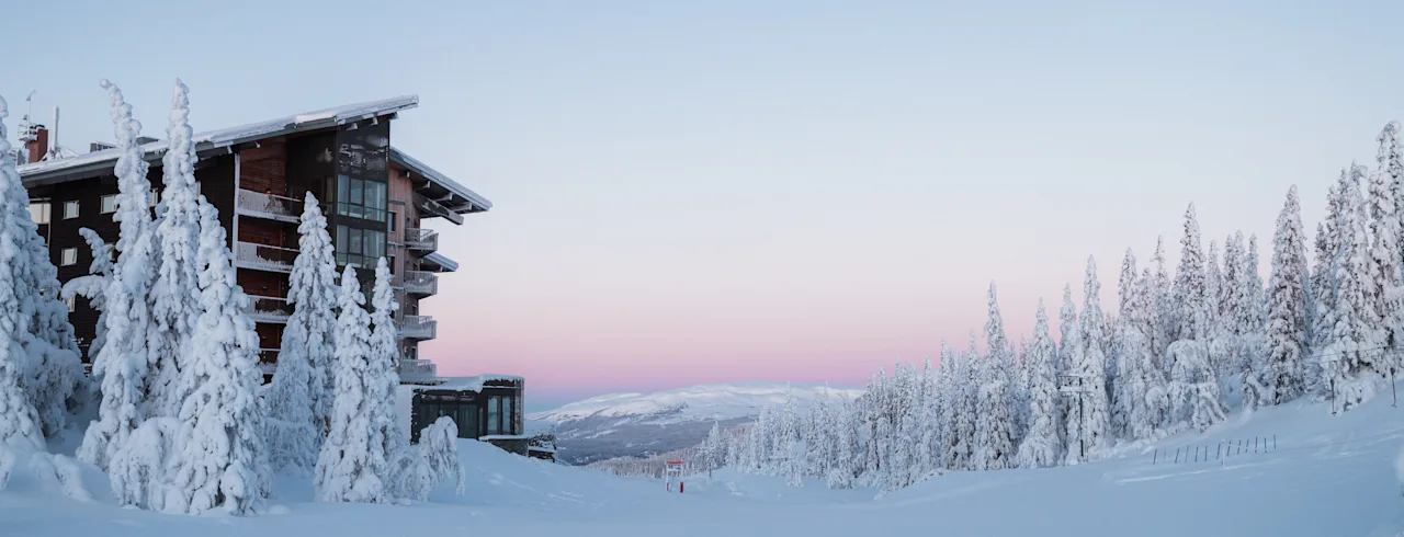Viihtyisä lomamökki lumipeitteisten mäntyjen keskellä rauhallisessa talvimaisemassa pehmeän vaaleanpunaisen ja sinisen taivaan alla.