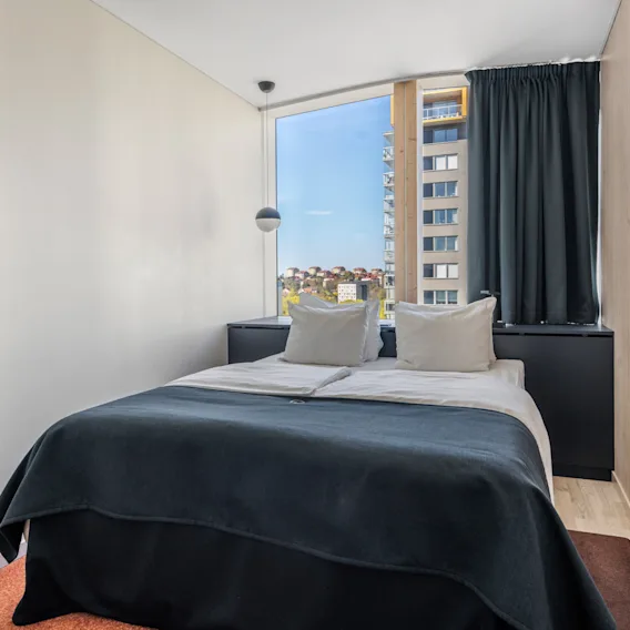 Bright hotel room featuring a made double bed in front of a large window with a city view.