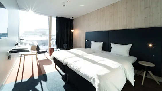 A bright hotel room with a large bed is neatly made; sunlight streams through a large window with dark curtains; a desk and two chairs are nearby.