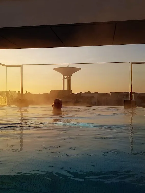 Quality Hotel™ Öresund – Rooftop Spa. En person kopplar av i en ångande takpool med en gyllene solnedgång över staden.