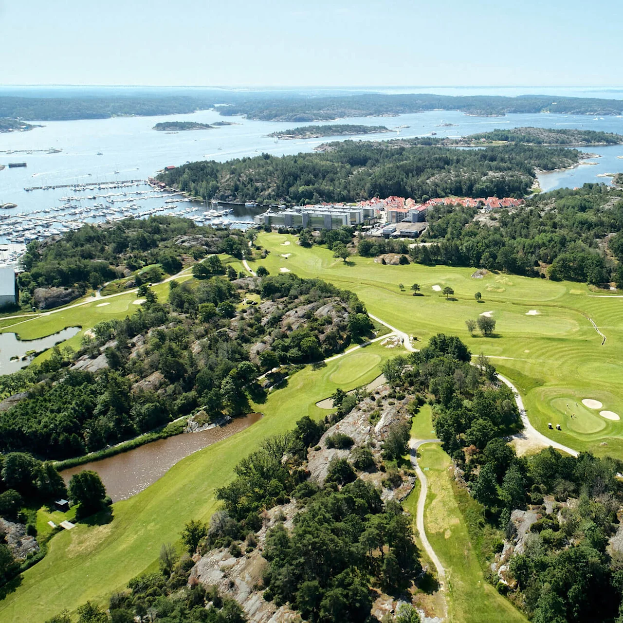En golfbane ligger i et steinete terreng, med flere sandbunkere og greener, omgitt av trær og med utsikt over et kystområde med båter og små øyer i det fjerne.