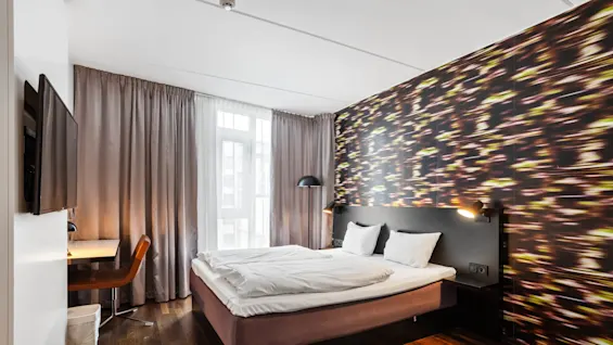 A modern hotel room features a double bed with white bedding, a patterned wall, a desk with a lamp, a chair, and a wall-mounted TV. Curtains cover large windows.