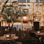 Bord med stearinlys er arrangerede foran en træhylde, der viser vaser og dekorationer, sat i en varm oplyst, hyggelig café eller restaurant, med et potte træ.