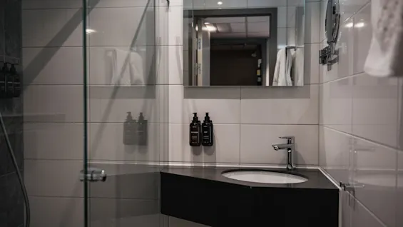 Et rent, moderne hotellbad med vask, speil og glassinnkapslet dusj. Veggmonterte dispensere er til stede.