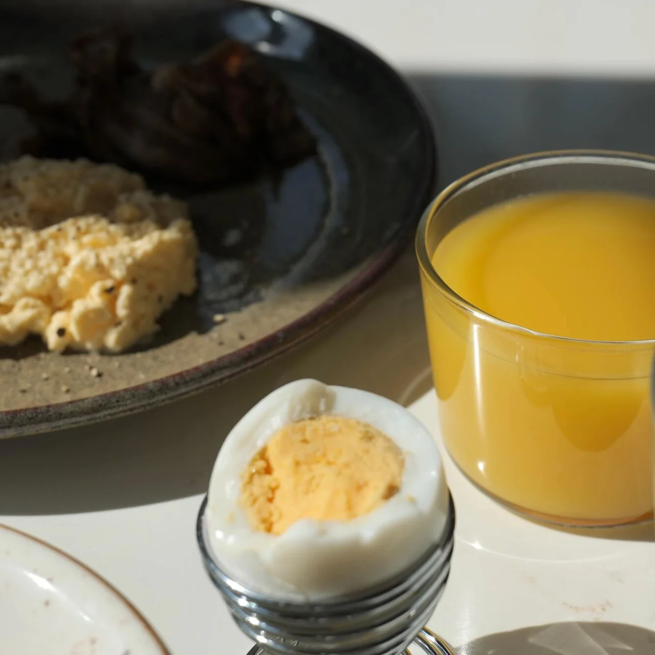 Et kogt æg står i en holder, med et glas appelsinjuice og en kop kaffe i nærheden på et solbeskinnet bord. En tallerken med røræg og kød ligger i baggrunden.