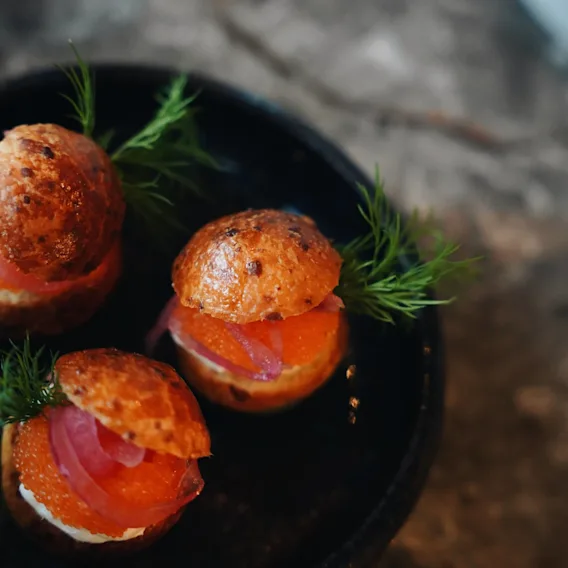 Små smörgåsar med orange fiskrom och rosa inlagd lök ligger i en mörk skål, garnerade med färsk dill, på en texturerad sten yta.