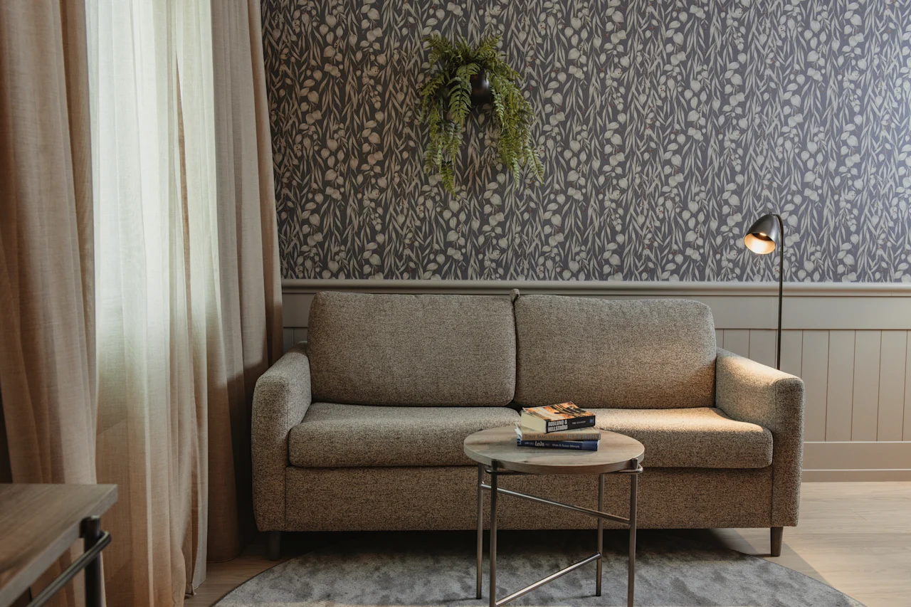 Et hyggeligt opholdsområde med en sofa, bøger på et bord og en gulvlampe, mod en mønstret væg.