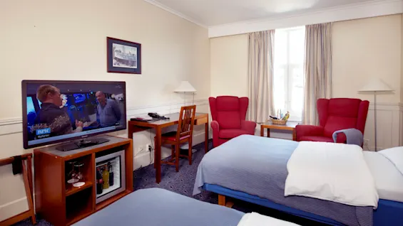 Ett hotellrum med två enkelsängar, en TV som visar en nyhetskanal, ett skrivbord, stolar och ett fönster med gardiner.