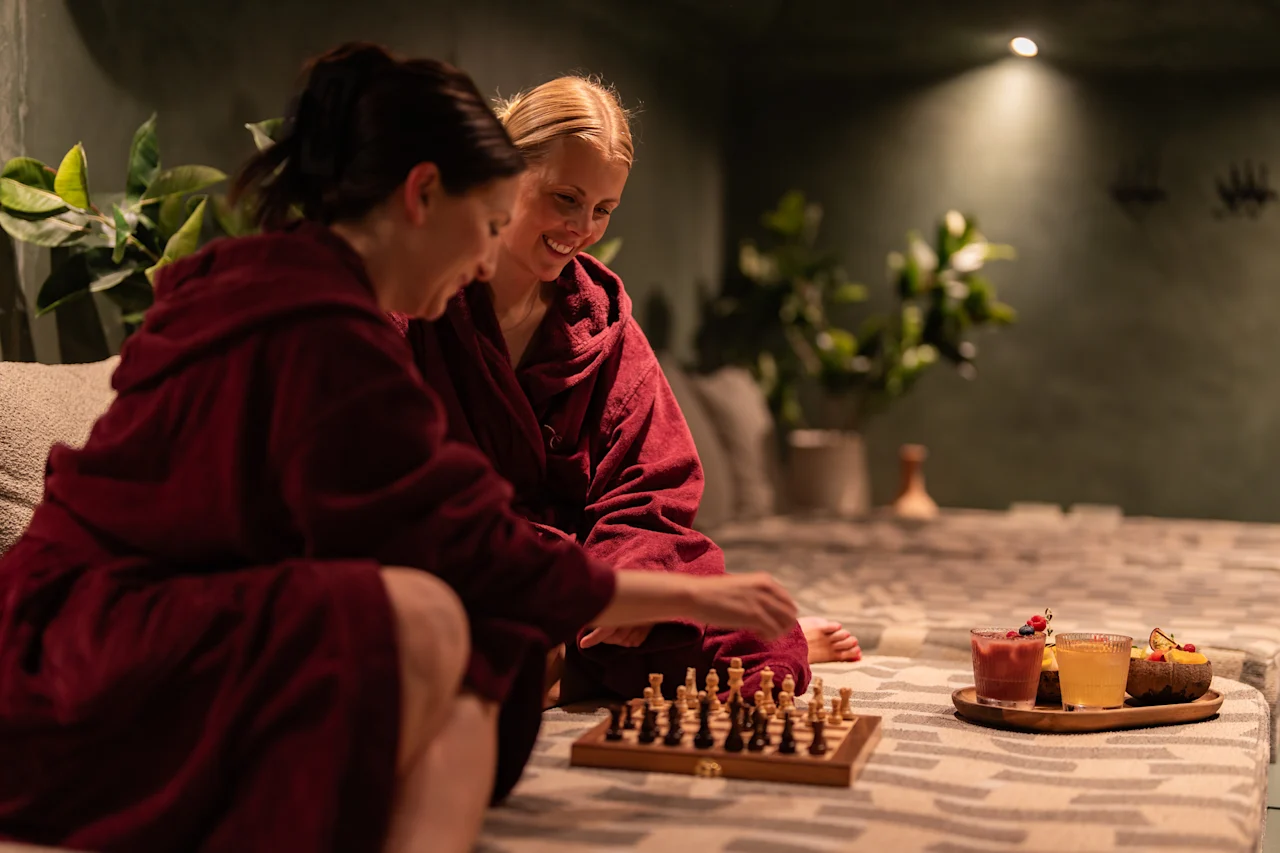 Kaksi kylpytakkiin pukeutunutta henkilöä nauttii shakkipelistä viihtyisässä, hämärästi valaistussa oleskelutilassa, virkistävien juomien ja välipalojen äärellä.