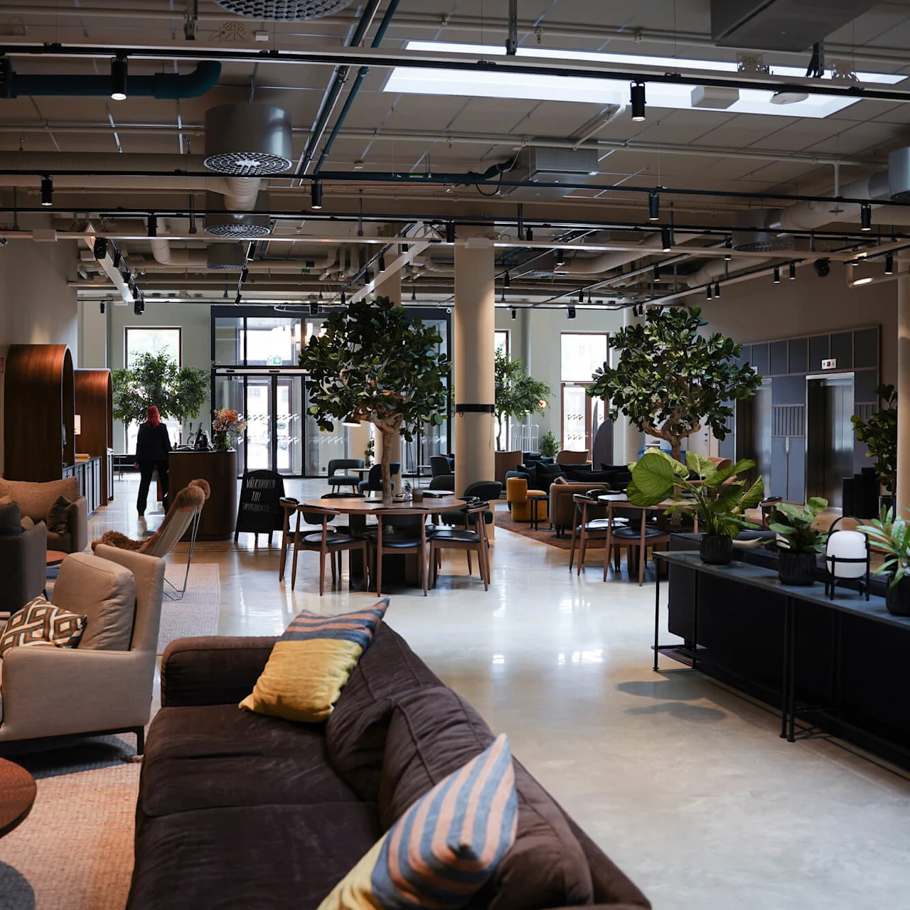 A modern lounge with sofas, chairs, plants, and decorative lighting. A person standing in the distance.
