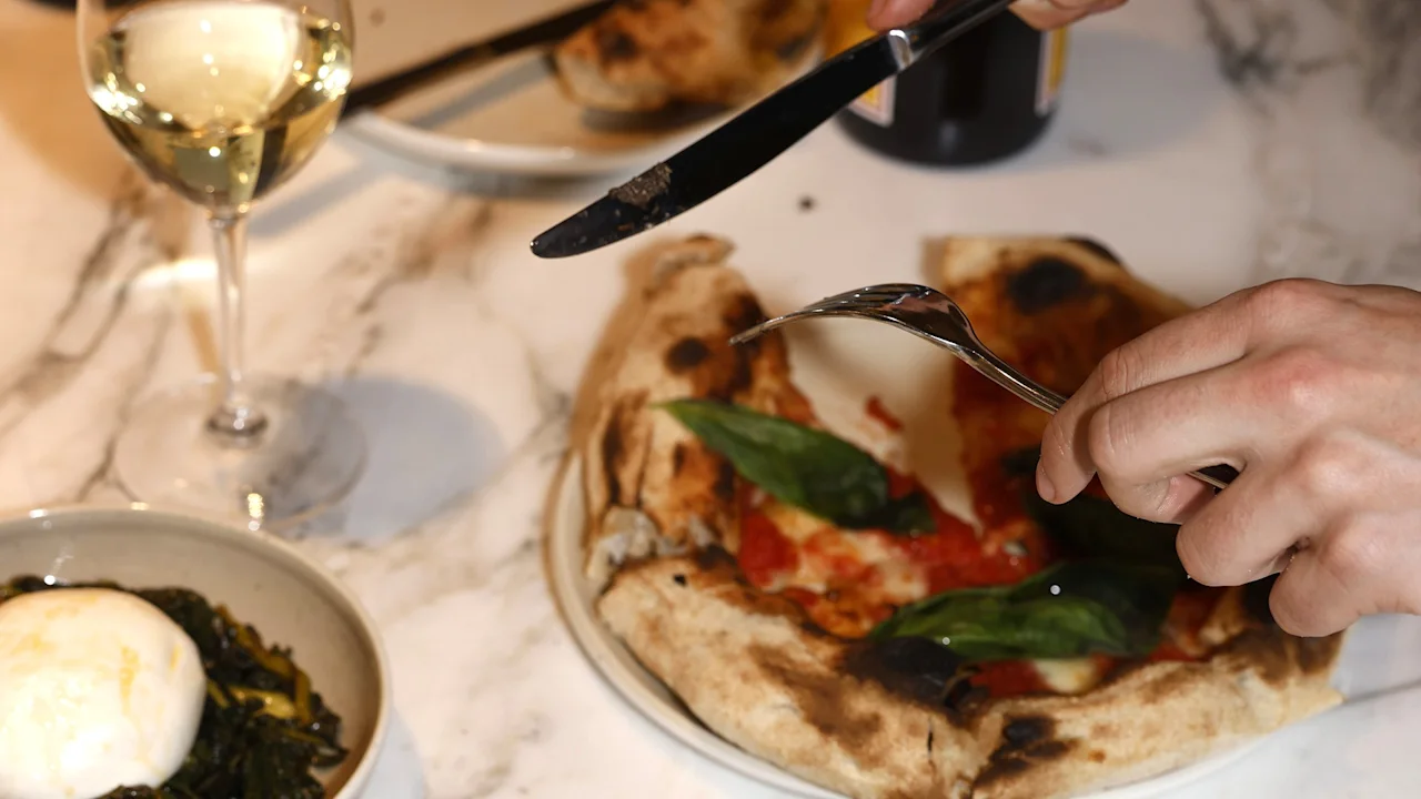 En person skærer en pizza med kniv og gaffel på et marmorbord. Nærved er der et glas hvidvin og en ret med ost og grønne blade.
