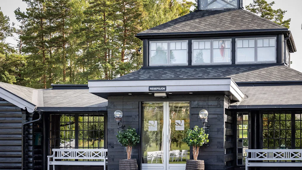 Ingången till en hotellreception med glasdörrar, flankeras av krukväxter och bänkar; omgivet av skogsområde. Text på dörren: "QUALITY HOTEL ENTRY", "RESEPSJON".