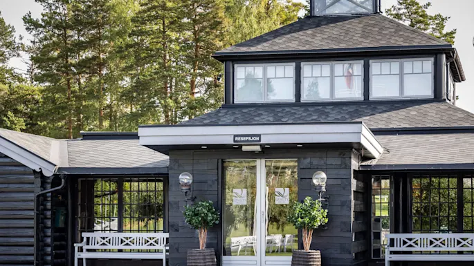 Entrance to a hotel reception with glass doors, flanked by potted plants and benches; surrounded by a wooded area. Text on door: "QUALITY HOTEL ENTRY", "RESEPSJON".