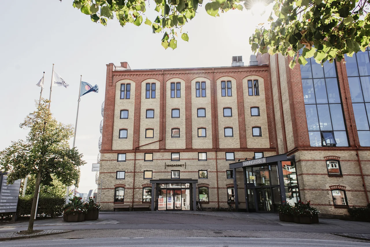 Suuri tiilirakennus, jossa modernit lasiosat ja sisäänkäynti. Liput ja puut kehystävät aurinkoista näkymää. MAGASINET, HAMNGATAN 9.