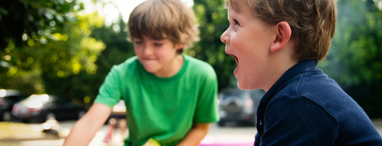 To barn leker gledelig i et rosa oppblåsbart basseng utendørs. Ett plasker vann mens det andre reagerer med åpen munn.
